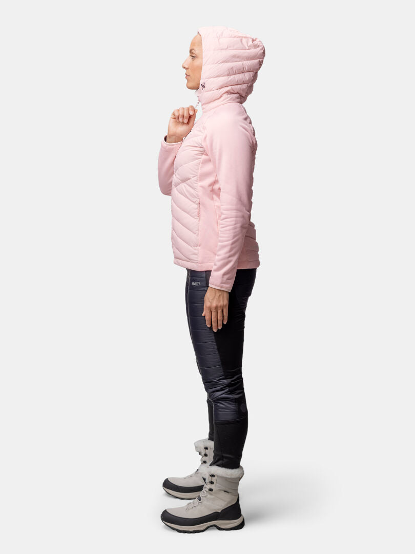Person i Halti Huippu Hybrid Dunjacka Dam (rosa), svarta byxor och grå stövlar mot en enfärgad bakgrund.