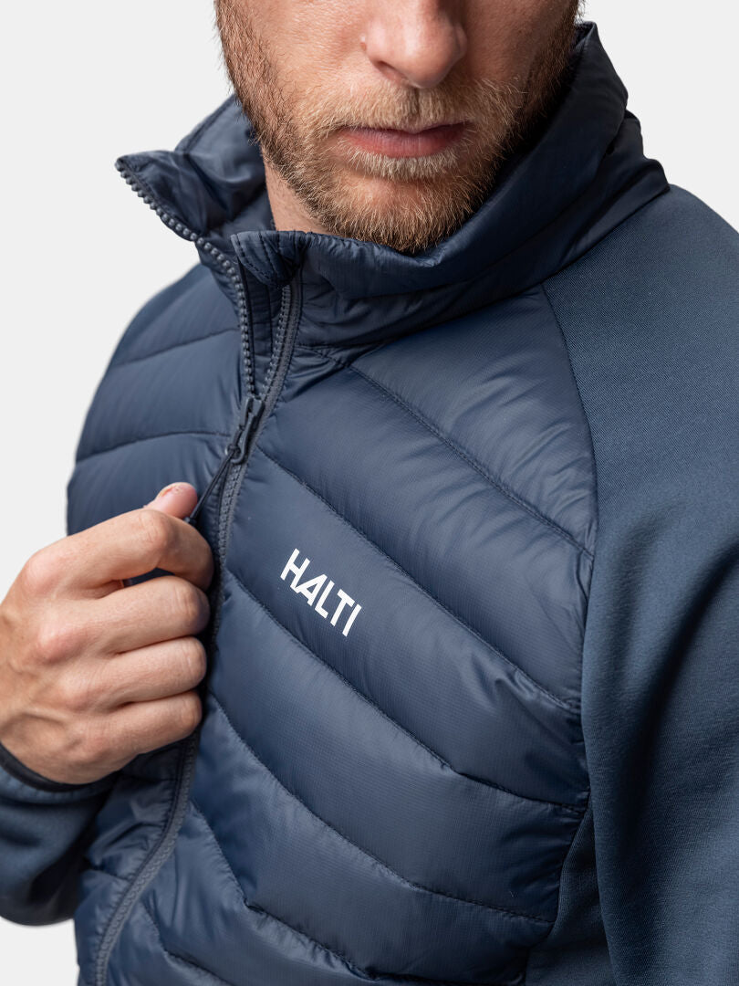 En man drar upp dragkedjan på en Halti Huippu Hybrid Dunjacka Herr i marinblått, som visas axel mot mun mot en enfärgad bakgrund.