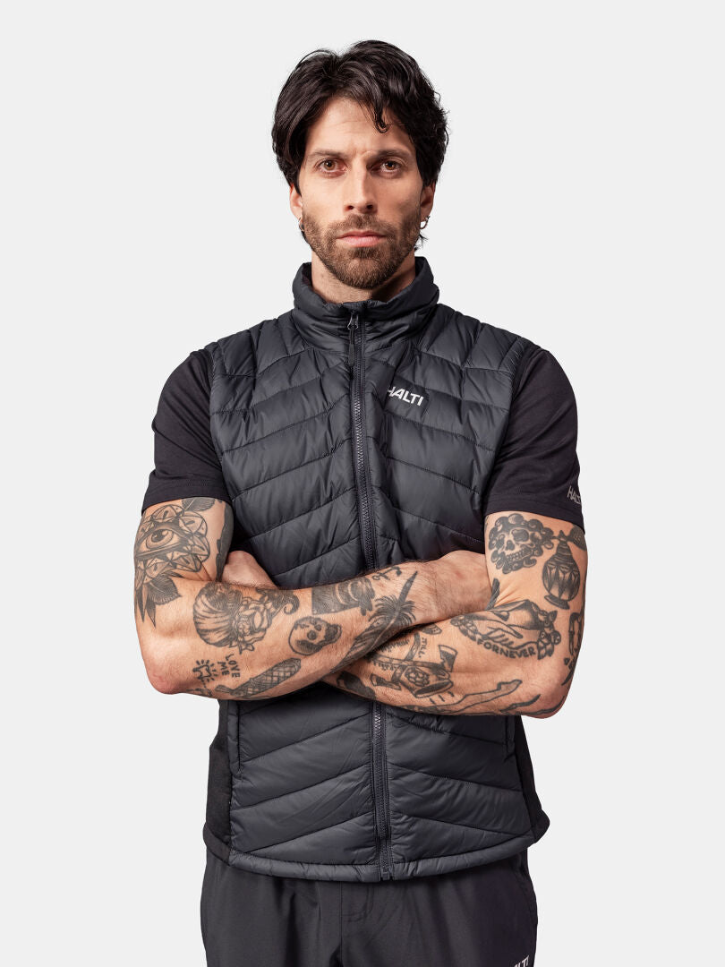 En tatuerad man bär en svart Halti Huippu Dunväst och en svart t-shirt med armarna i kors.