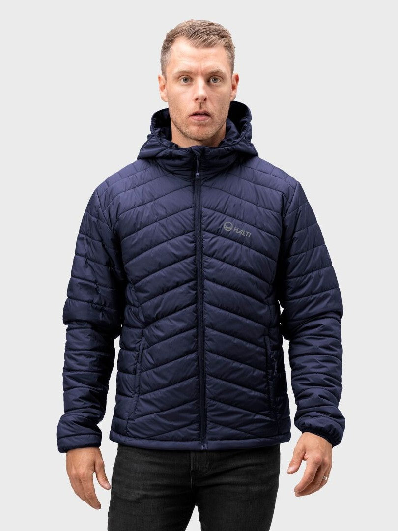 En man bär en Halti Komu Lätt Vadderad Jacka Herr med huva och svarta byxor, stående mot en enfärgad bakgrund.