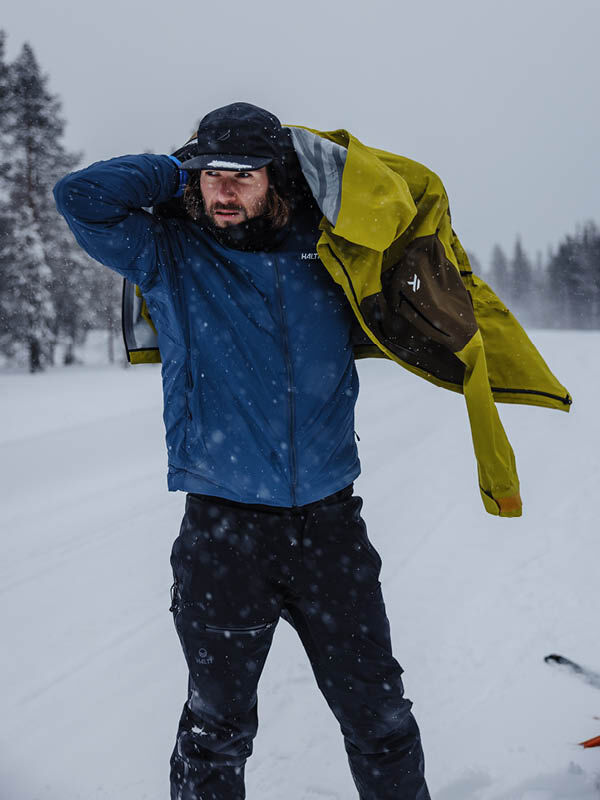 En vinterklädd man tar på sig en gul Halti Rizle 3L DX Skidjacka Unisex utomhus med snöiga träd bakom sig.