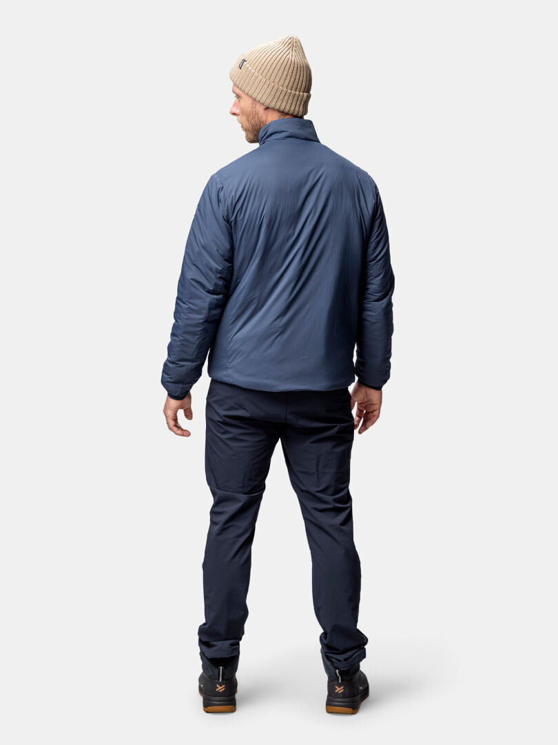 En man i beige stickad mössa och blå Halti Haave Insulation Primaloft Jacka Herr står mot en ljus bakgrund.