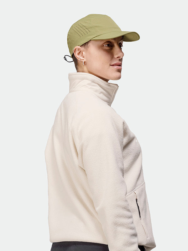 Person i ljusbeige jacka och olivgröna Halti Aero II Keps stående snett mot en enfärgad ljus bakgrund.