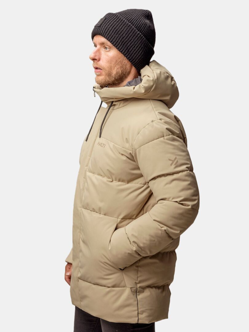 En man i en beige dunjacka och en Halti Huurre-mössa, som står i profil mot en enfärgad ljus bakgrund.