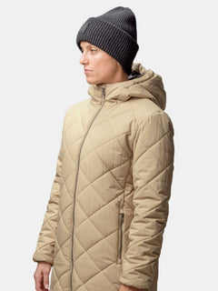 En person i beige quiltad kappa och Halti Huurre Mössa står mot en ljus bakgrund.