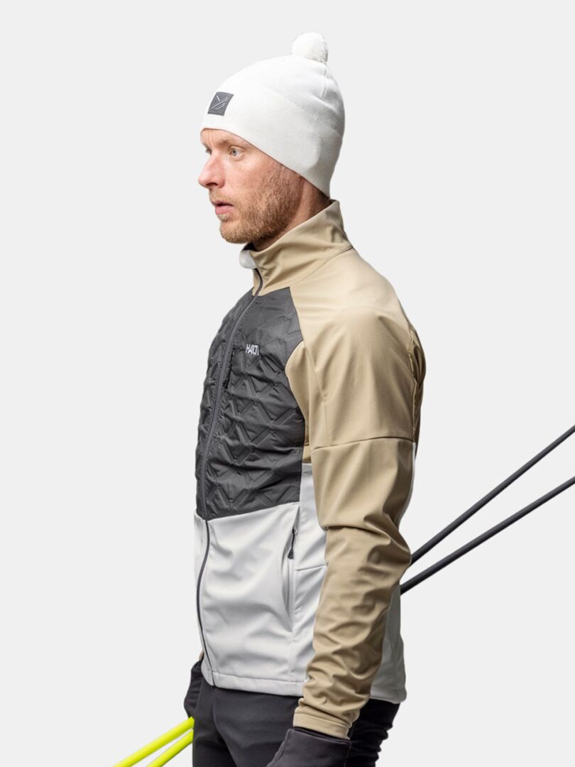 En man i beige, grå och svart jacka bär en Halti Pilke Mössa och håller i skidstavarna, redo för längdskidåkning.