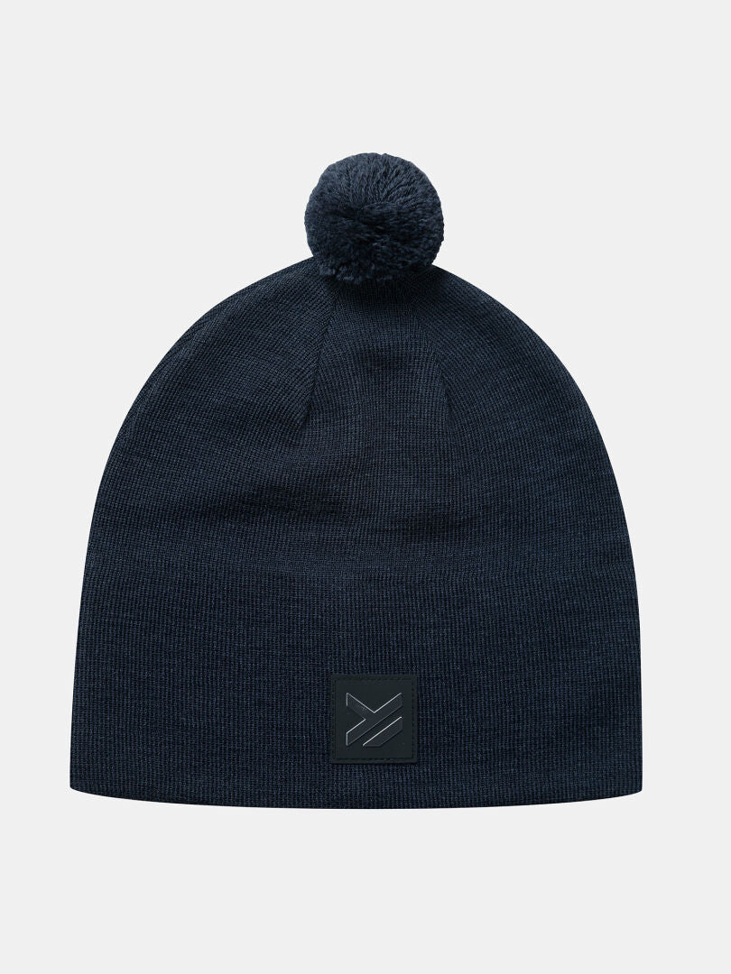 Halti Pilke Mössa: Mörkblå stickad mössa med pompon och svart patch, perfekt för vintersportäventyr.