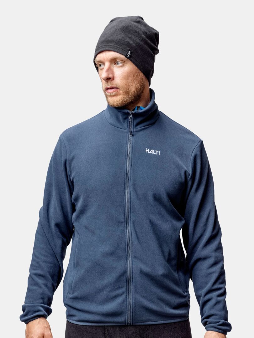 Man i blå Halti zip-up jacka och Trone Fleece Mössa står redo för vinterträning mot en vit bakgrund.