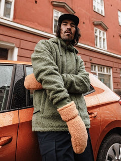 En man i grön fleecejacka och Halti Lykky Fleecevantar står vid en orange bil nära en röd byggnad.