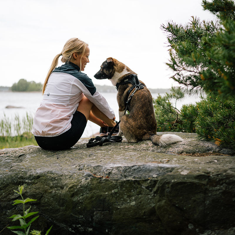 En kvinna i Halti Urbanite Dam Träningsjacka sitter vid vattnet med sin fastspända hund, omgiven av träd.