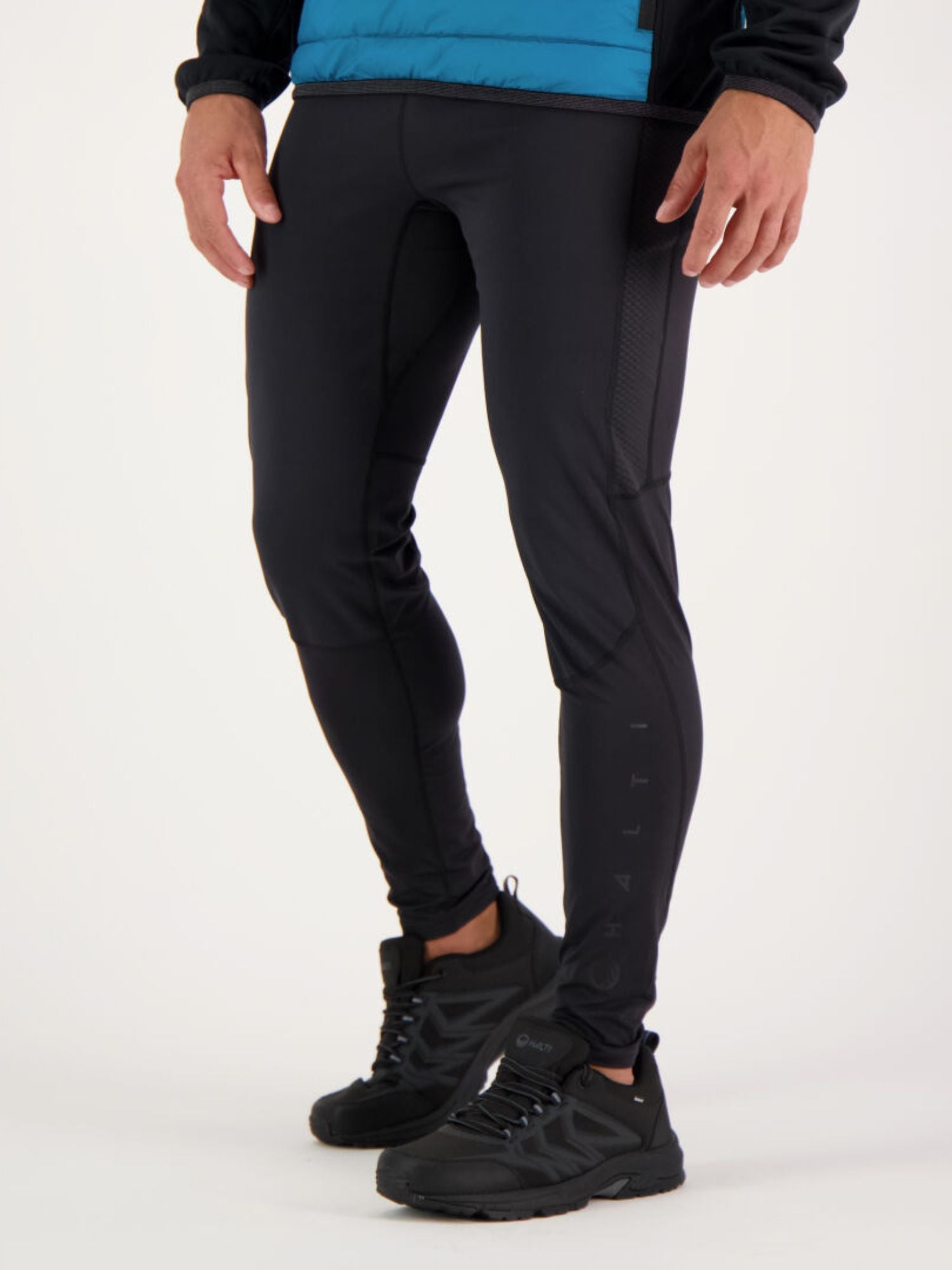 En person i Halti Suunta Herr Hybridtights, svarta sneakers och en blå jacka står mot en enfärgad bakgrund.