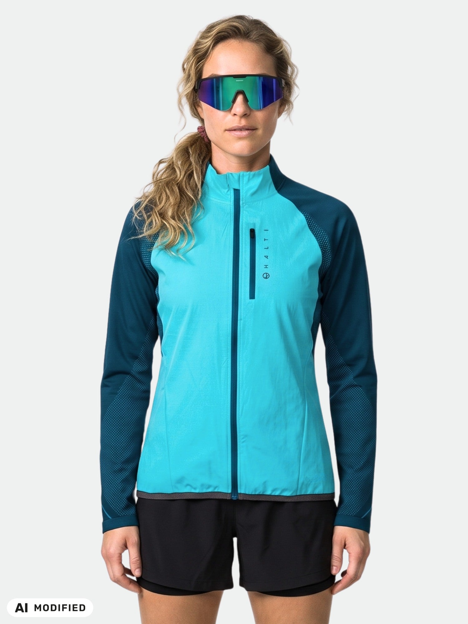 En person klädd i en Halti Tracks Dam Seamless Jacka och svarta shorts, med reflekterande solglasögon, står framför en enfärgad bakgrund.