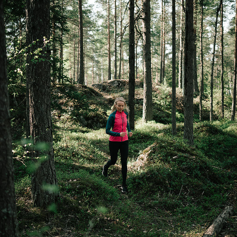 Någon i en rosa Halti Tracks Dam Seamless Jacka joggar på en skogsstig med gröna träd och mossig mark.