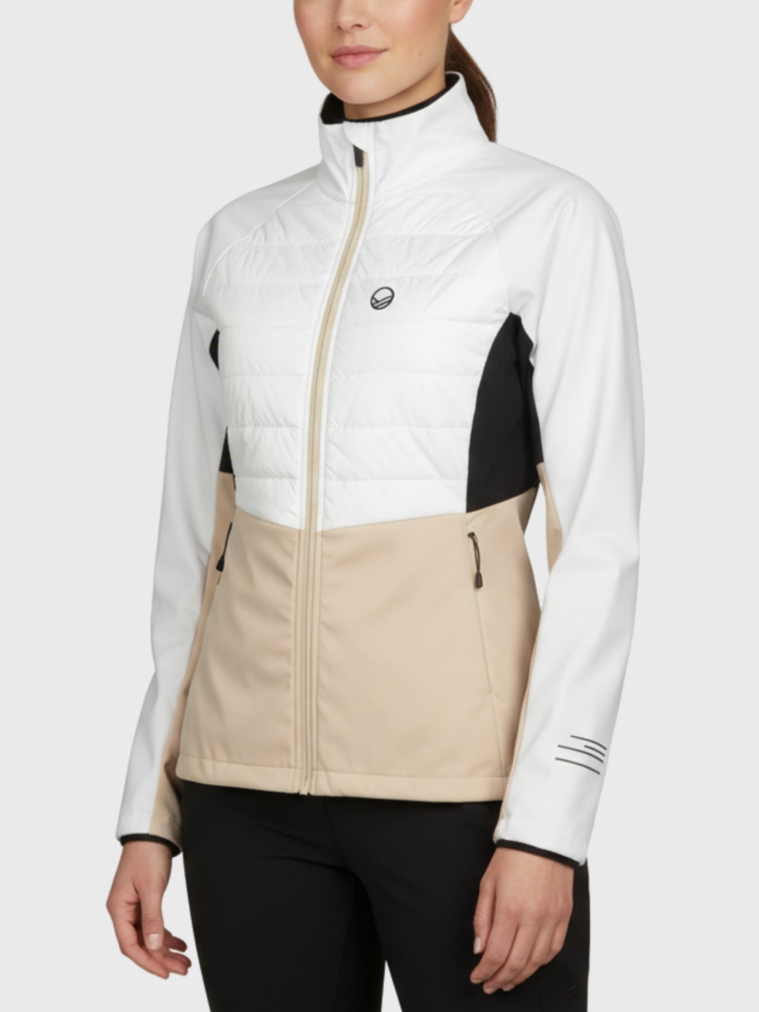 En kvinna bär Halti Vinha Hybrid Längdskidjacka Dam med vita, beige och svarta paneler, dragkedja fram och fickor.