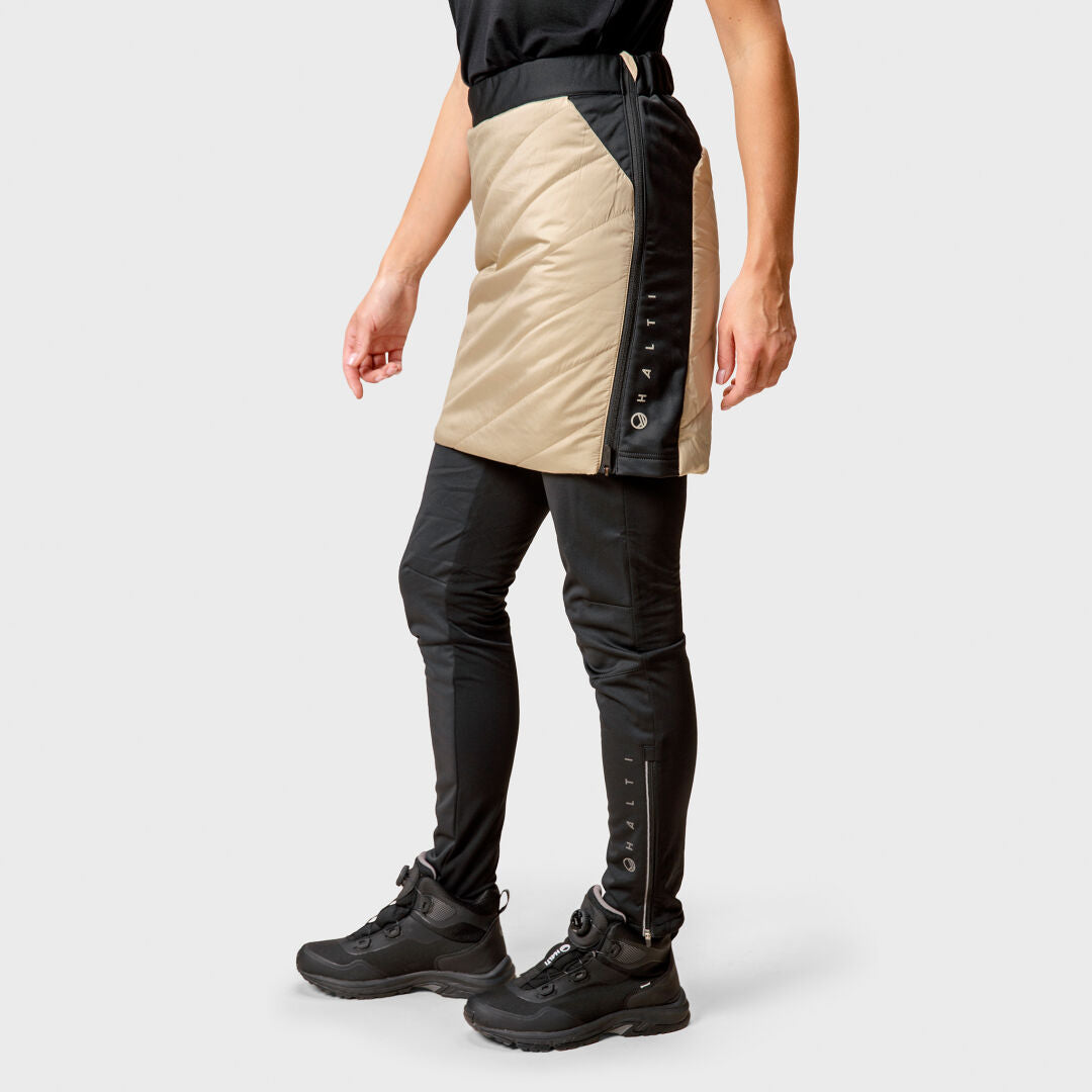 En person bär Halti Hanki Warm Hybrid Kjol Dam-byxor och vandringsskor, stående mot en enfärgad bakgrund.