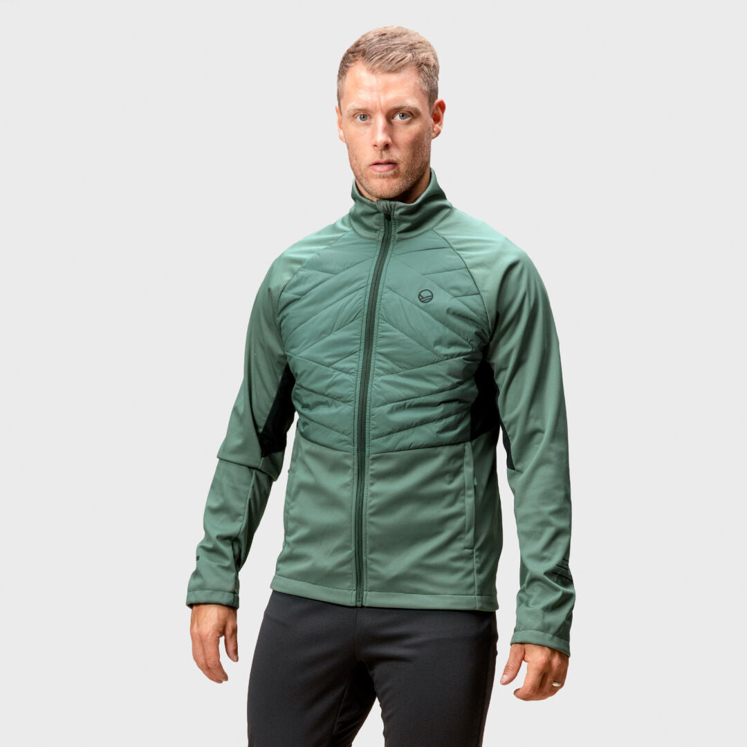 En man i en grön Halti Vinha Hybrid Längdskidjacka Herr med Stormwall® står vänd framåt mot en ljusgrå bakgrund.