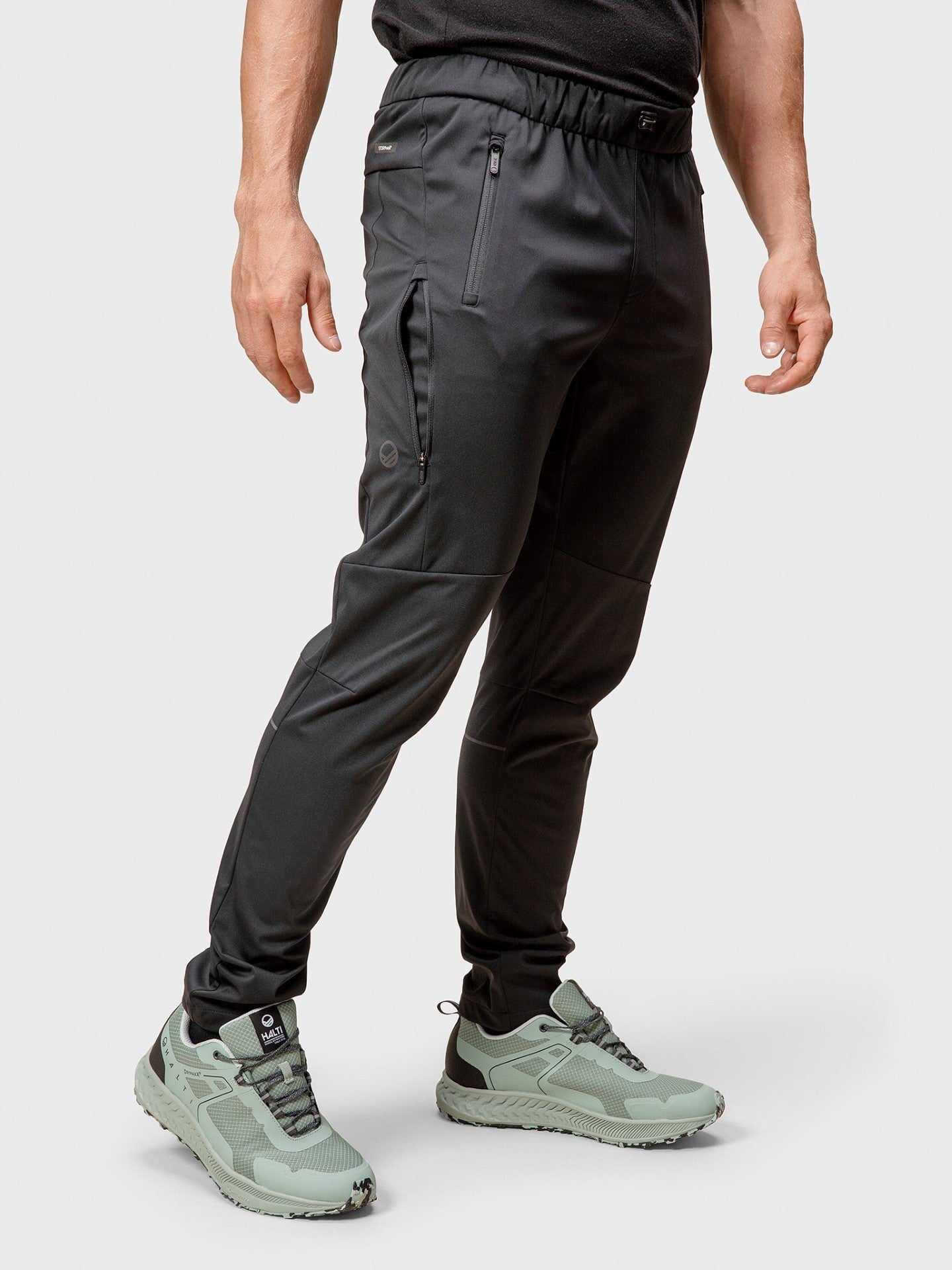 En person i Halti Exhale Stormwall Byxor Herr och ljusgröna sneakers står mot en vanlig ljus bakgrund.
