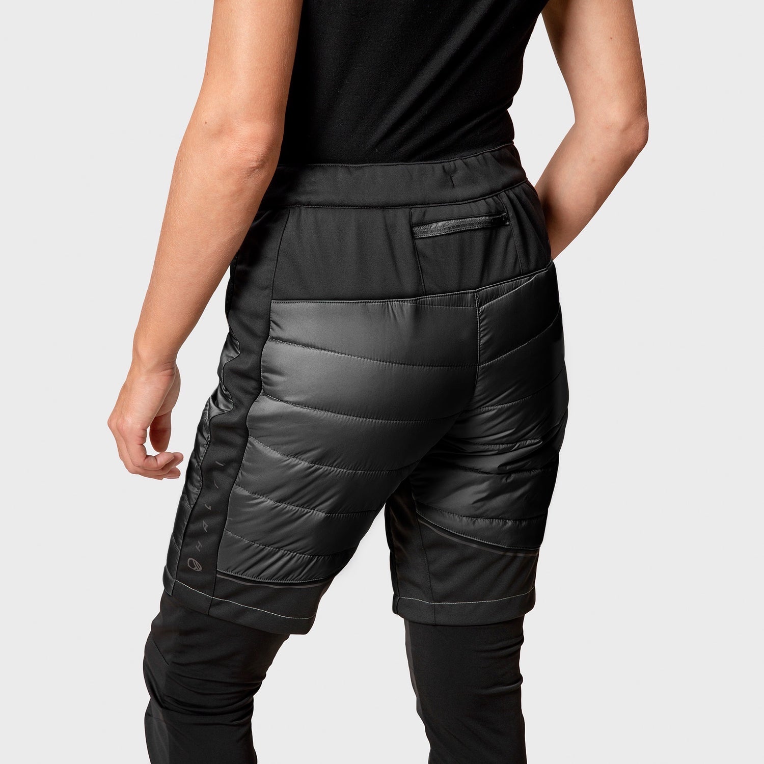 Person i Hanki Warm Hybrid Shorts Dam från Halti över svarta byxor, sedd bakifrån mot ljus bakgrund.
