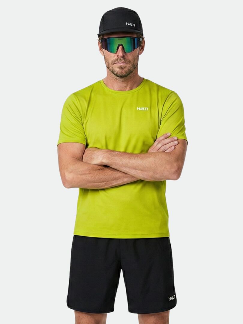 Man i limegrön Halti Salves T-skjorta Herr, svarta shorts och keps står med armarna i kors mot enfärgad bakgrund.