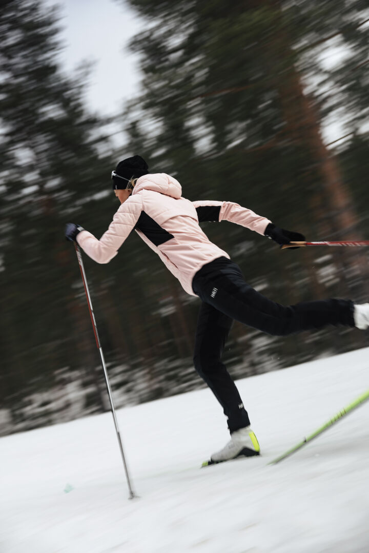 En person i Halti Hanki 2 Hybrid Längdskidbyxor Dam längdskidor snabbt på snö med en skog i bakgrunden.