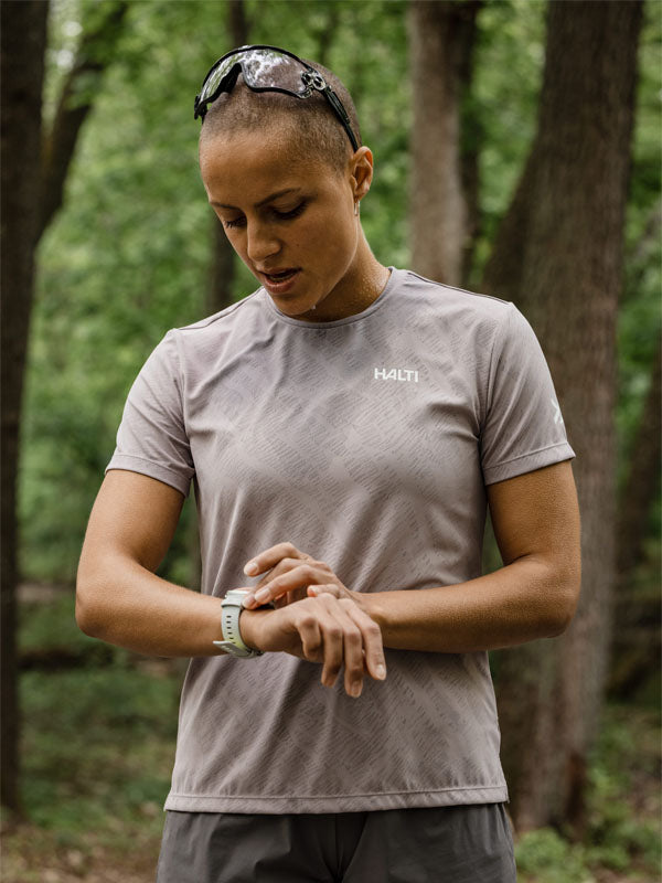 En person i en grå Halti Oulanka T-skjorta Dam kontrollerar en smartklocka när han står i ett skogsområde.