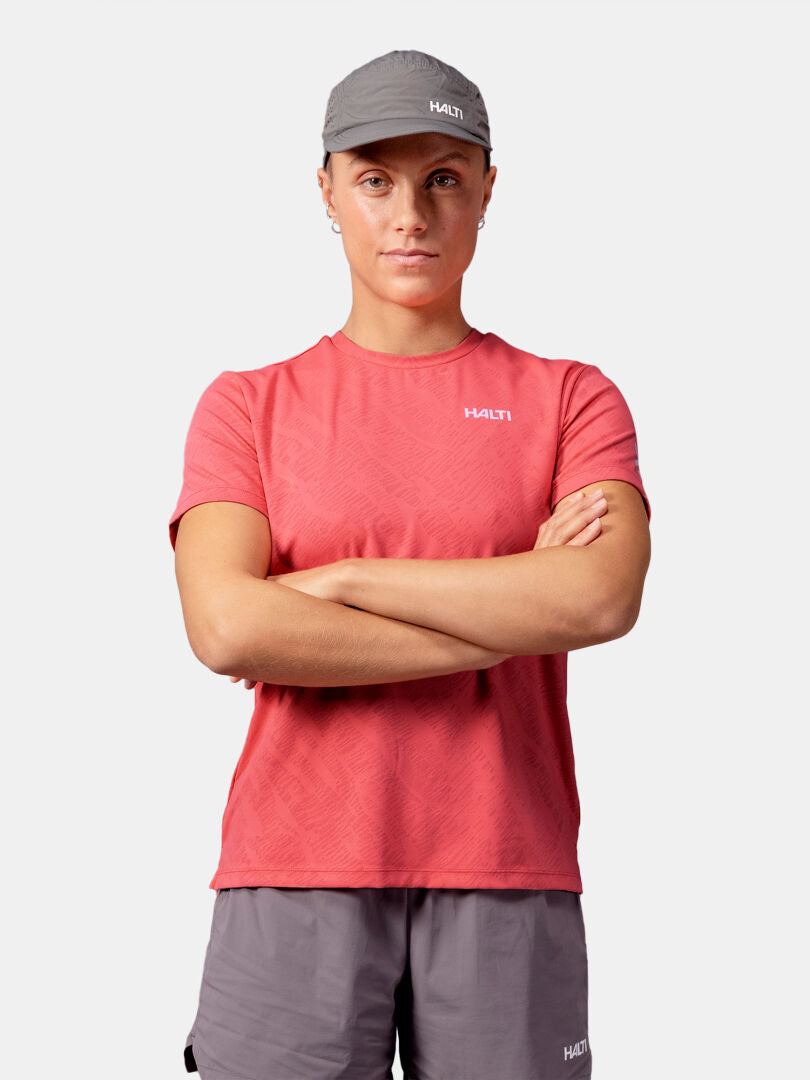 Person i röd Halti Oulanka T-skjorta Dam, grå shorts och grå keps står med armarna i kors mot en enfärgad bakgrund.