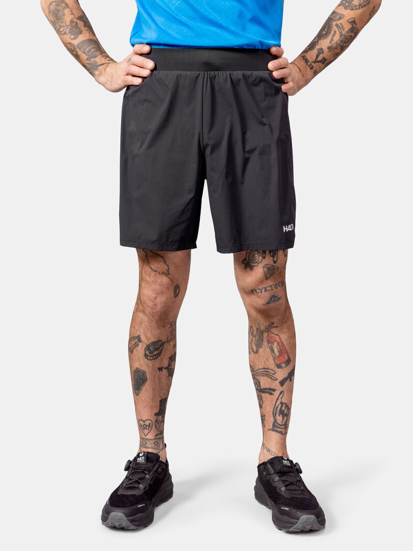 Person i Halti Uurto Shorts Herr, blå skjorta, svarta skor; står med händerna på höfterna, tatuerade ben synliga.