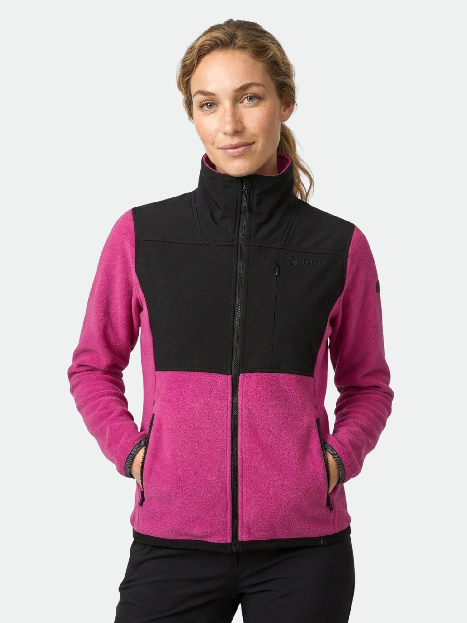 En kvinna i en rosa och svart Halti Pioneering Fleecejacka Dam står med händerna i fickorna mot en enfärgad bakgrund.
