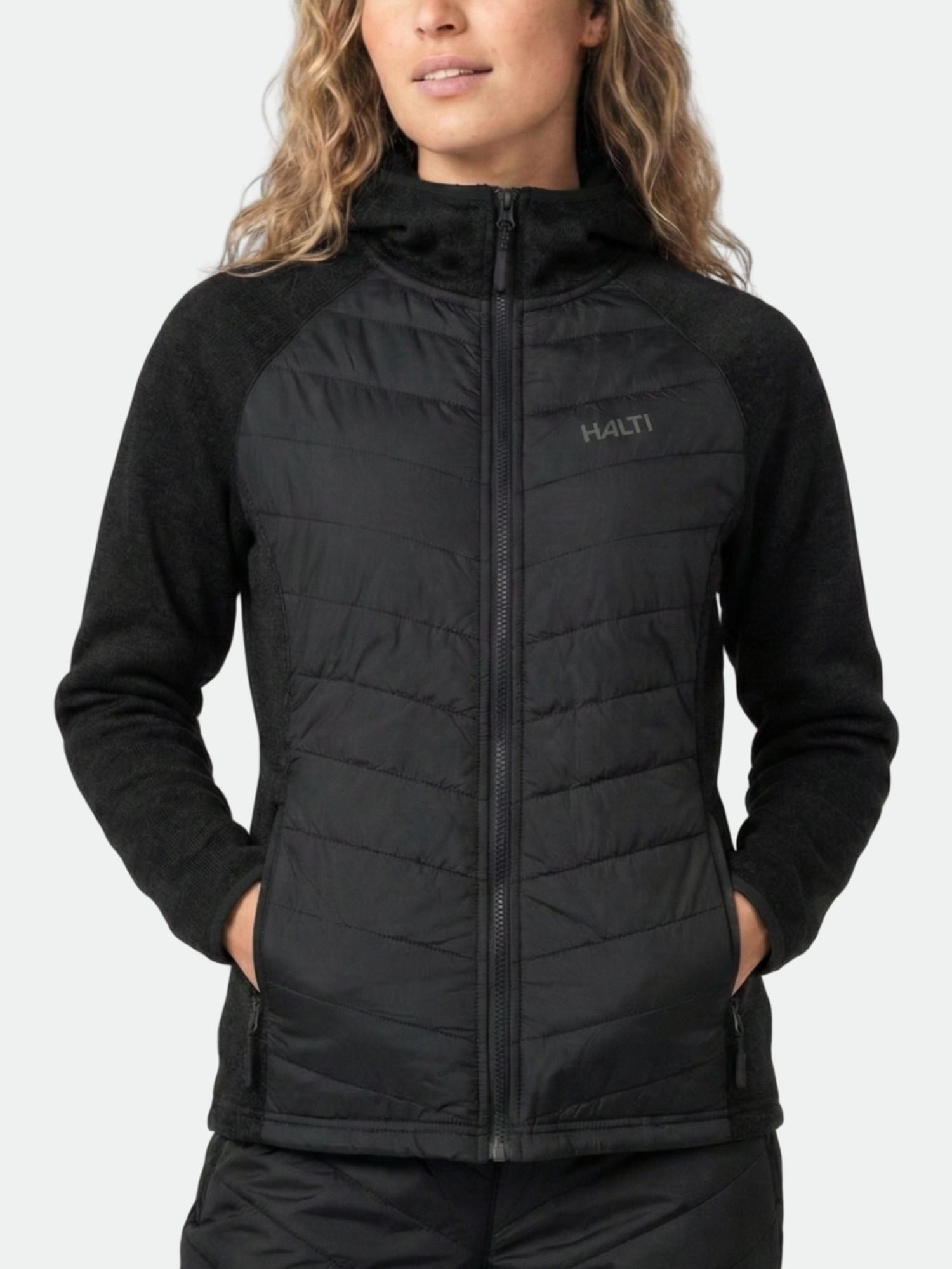 En person klädd i en svart Halti Streams Hybrid Mellanlager Dam-jacka med händerna i fickorna står mot en enfärgad bakgrund.
