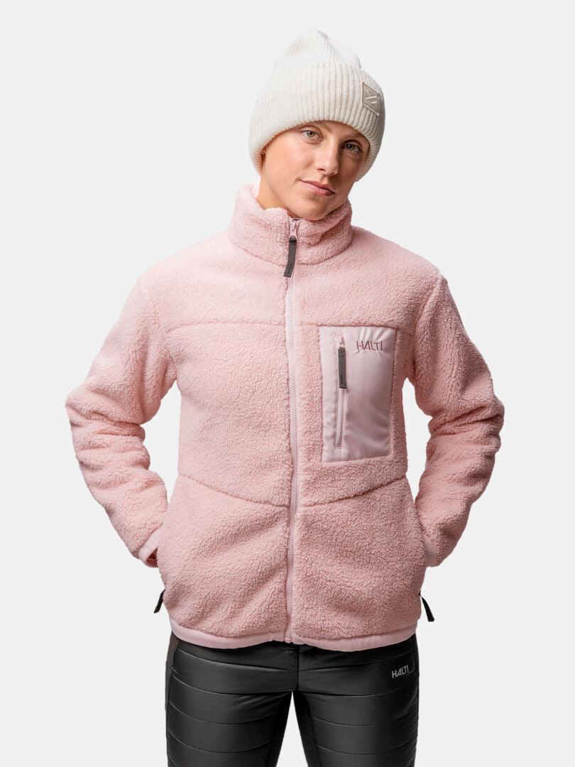 Personen bär en Halti Lumista Polar Fleecejacka för dam i rosa, en vit stickad mössa och svarta byxor, med händerna i jackfickorna.