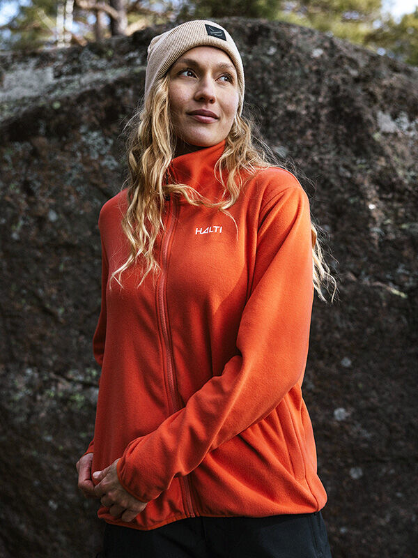 Kvinna i orange Halti Rockmoon Fleecejacka Dam och beige mössa står utomhus vid en stor sten.