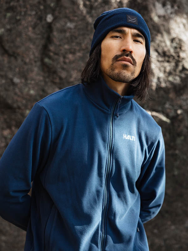 En person med långt hår och mustasch bär en Halti Rockmoon Fleecejacka Herr och mössa, utomhus vid en sten.