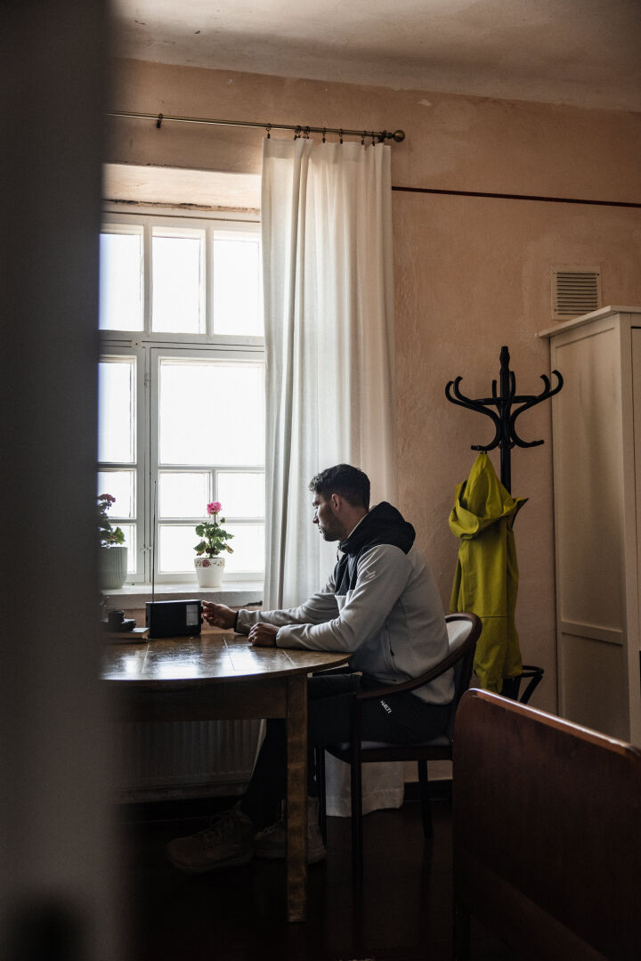 En man sitter vid ett fönster med krukväxter, och hans Halti Rockmoon Fleecehoodie Herr hänger på klädhängaren bredvid honom.