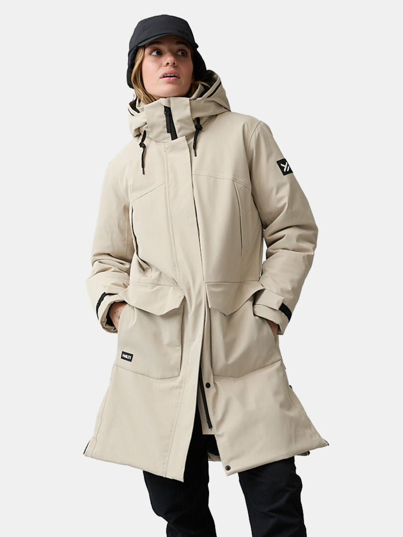 En person klädd i en beige Halti Kallio DX Parka Dam med huva, svart keps och byxor, framför en enfärgad ljus bakgrund.