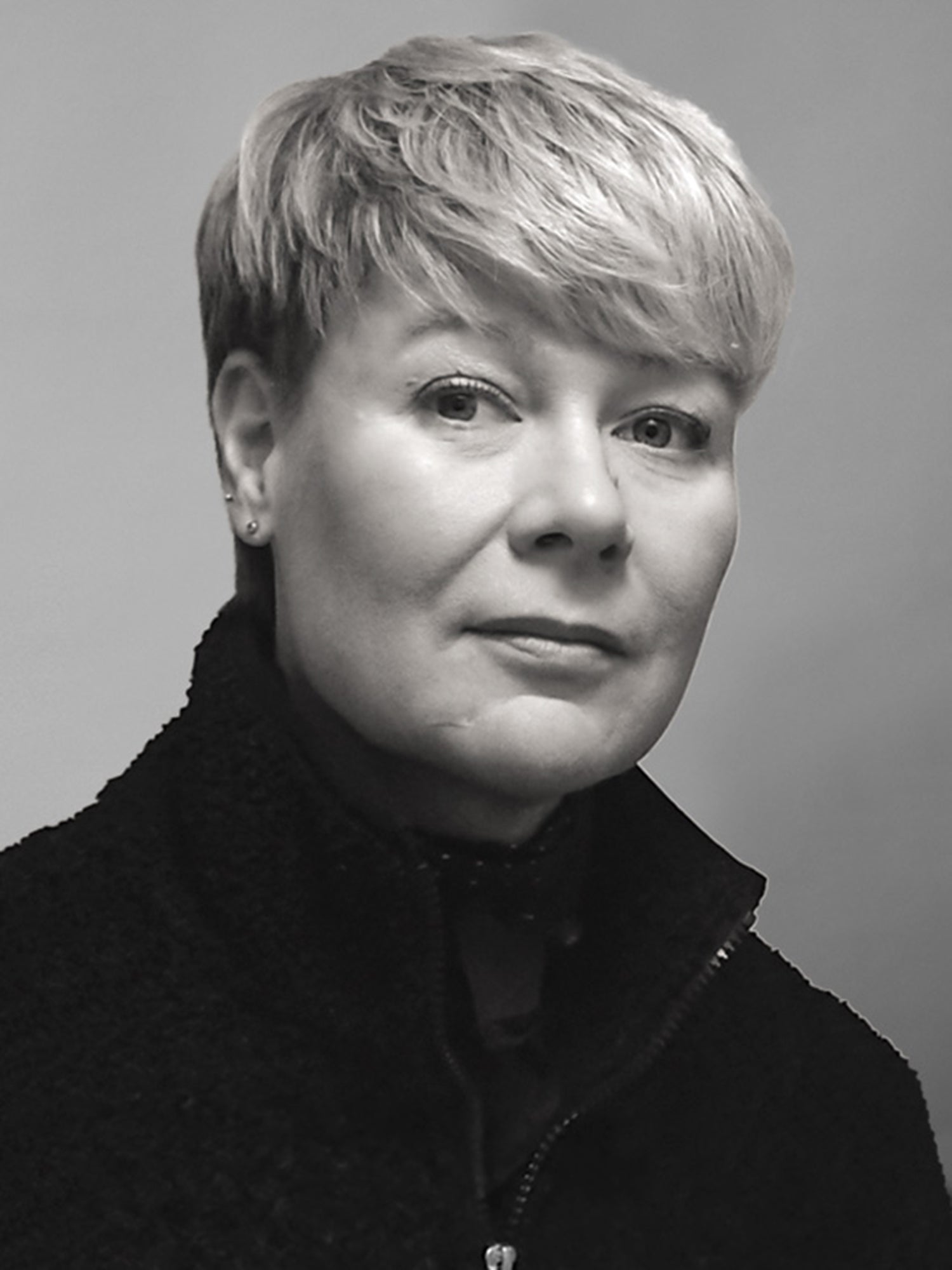 En person med kort, ljust hår och mörk jacka med dragkedja tittar in i kameran mot en enfärgad bakgrund.