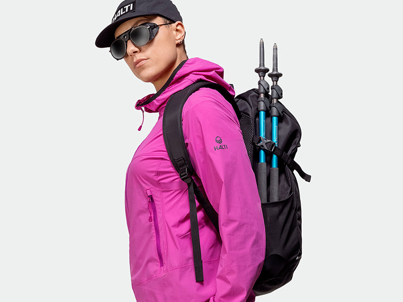 En person i rosa jacka och svart keps bär solglasögon och en ryggsäck med vandringsstavar fästa på sidan.