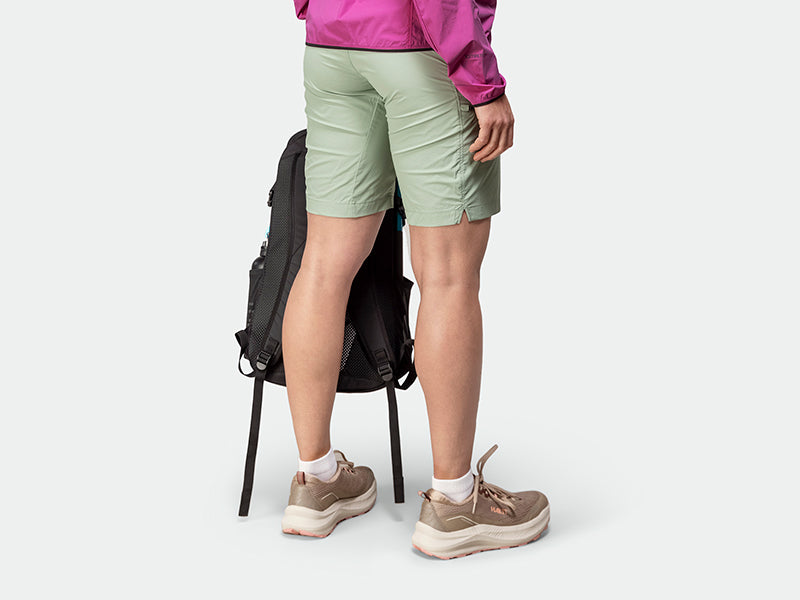 Person klädd i gröna shorts, bruna sneakers och rosa jacka, står bredvid en svart ryggsäck mot en enfärgad bakgrund.