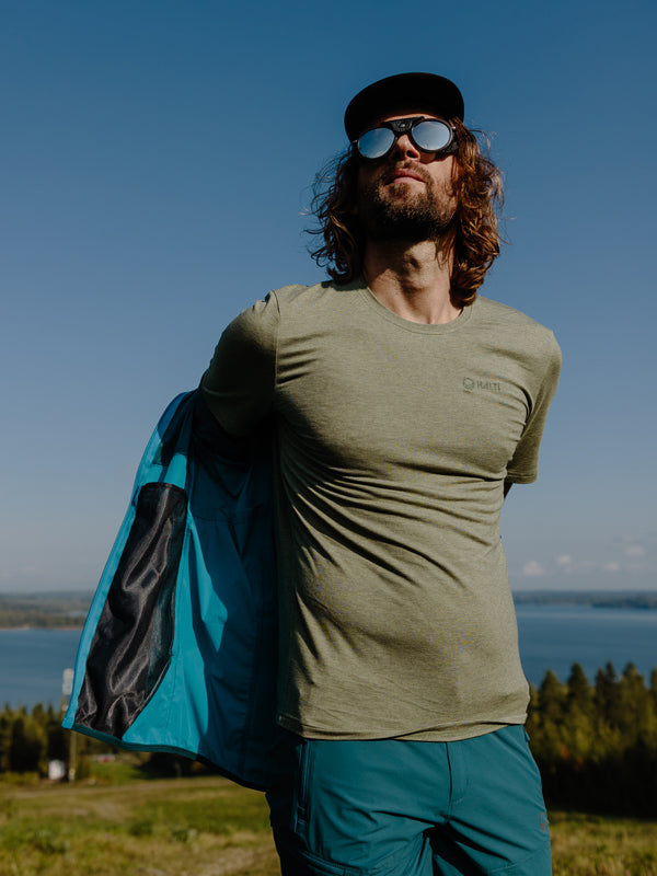 En man med solglasögon och keps står utomhus och tar på sig en blå jacka över en grön t-shirt.