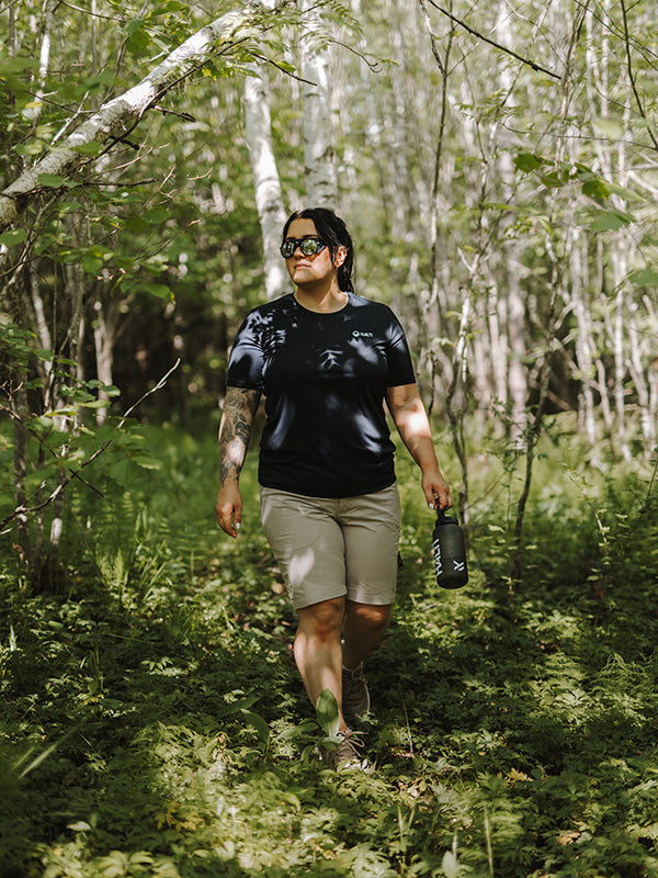 En person går genom en solbelyst skogsstig, klädd i solglasögon, mörk skjorta, shorts och med en vattenflaska i handen.