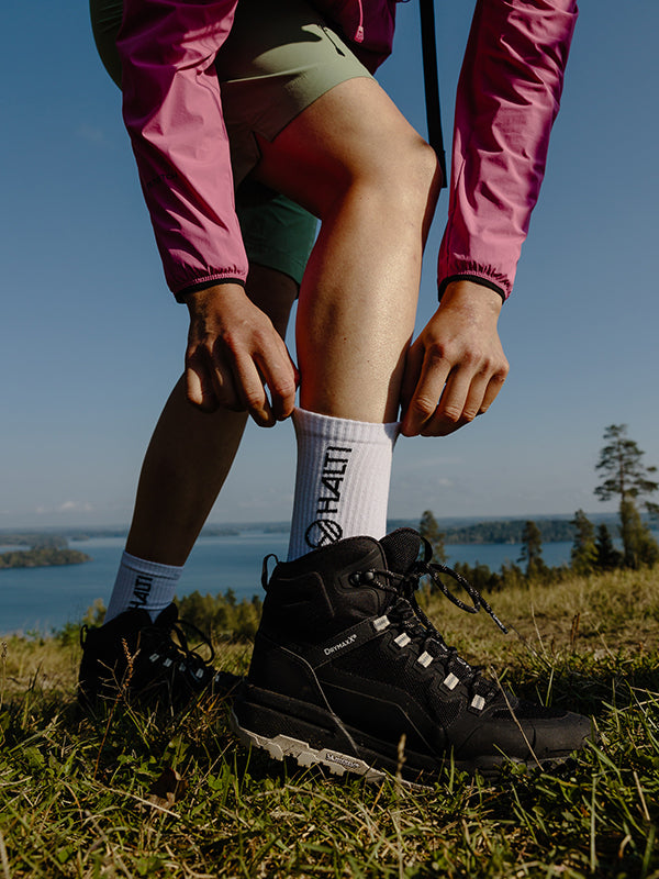 Person i rosa jacka, shorts med vit strumpa och svarta vandringskängor utomhus nära en sjö och träd.