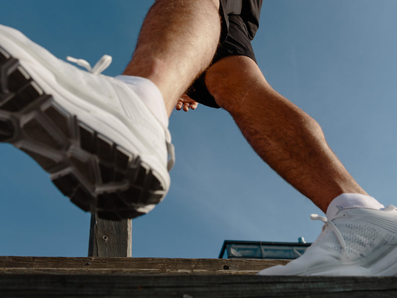Lågvinkelbild av en person i vita sneakers och shorts som går utomhus på en träyta mot en blå himmel.