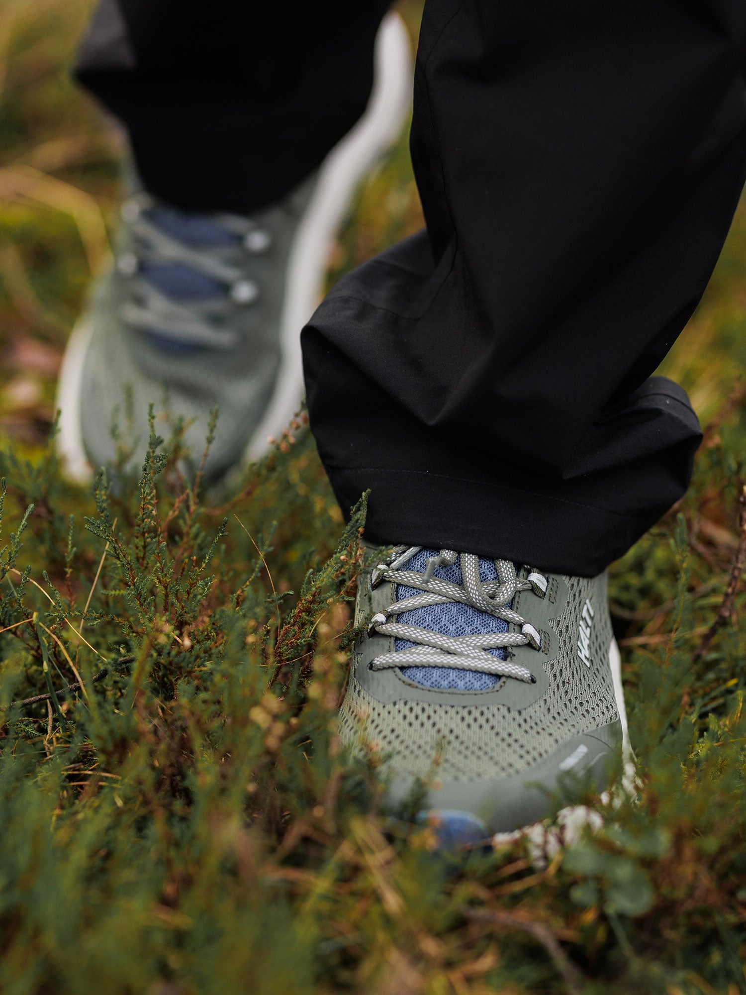 Person klädd i Halti Rapid 2 Sneaker Herr och svarta byxor, stående på gräs och gröna växter utomhus.