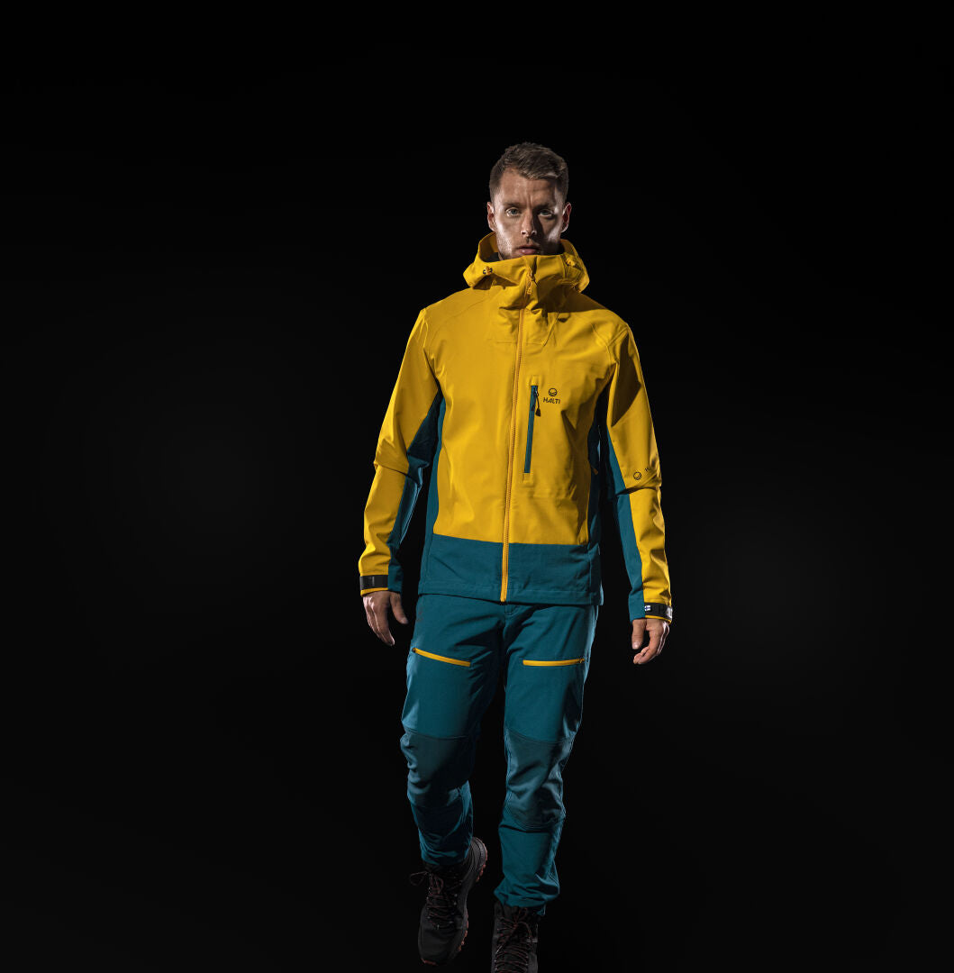 En man bär Halti Pallas II Herr Warm X-Stretch Byxor och en gul luvjacka, redo för friluftsliv mot svart.