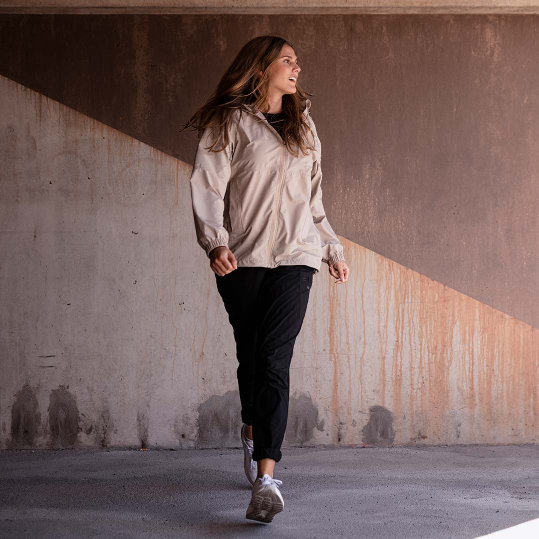 En kvinna i en beige Halti Pallas Dam Jogger Lite Jacka går inomhus längs en vägg med diagonala ljusa och mörka partier.
