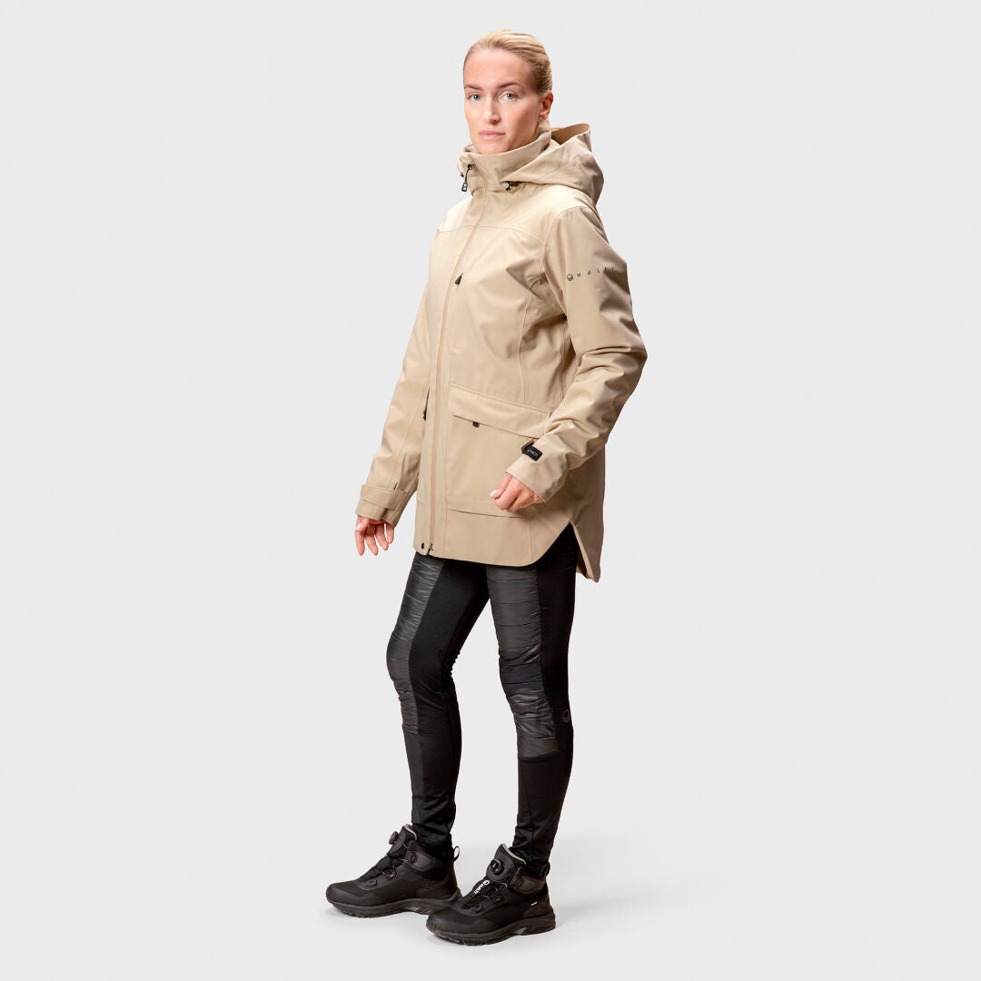 En person i en beige Halti Olhava DrymaxX Parka Dam, svarta byxor och stövlar står mot en ljusgrå bakgrund.