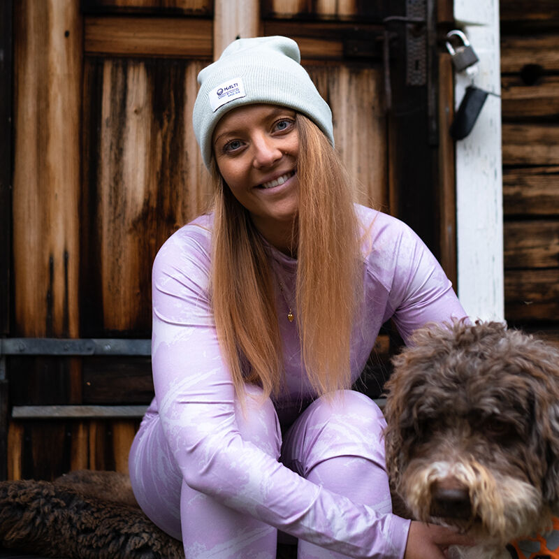 En kvinna i Halti Windfire Underställsset-dammen och en grå mössa ler utanför med sin bruna hund.